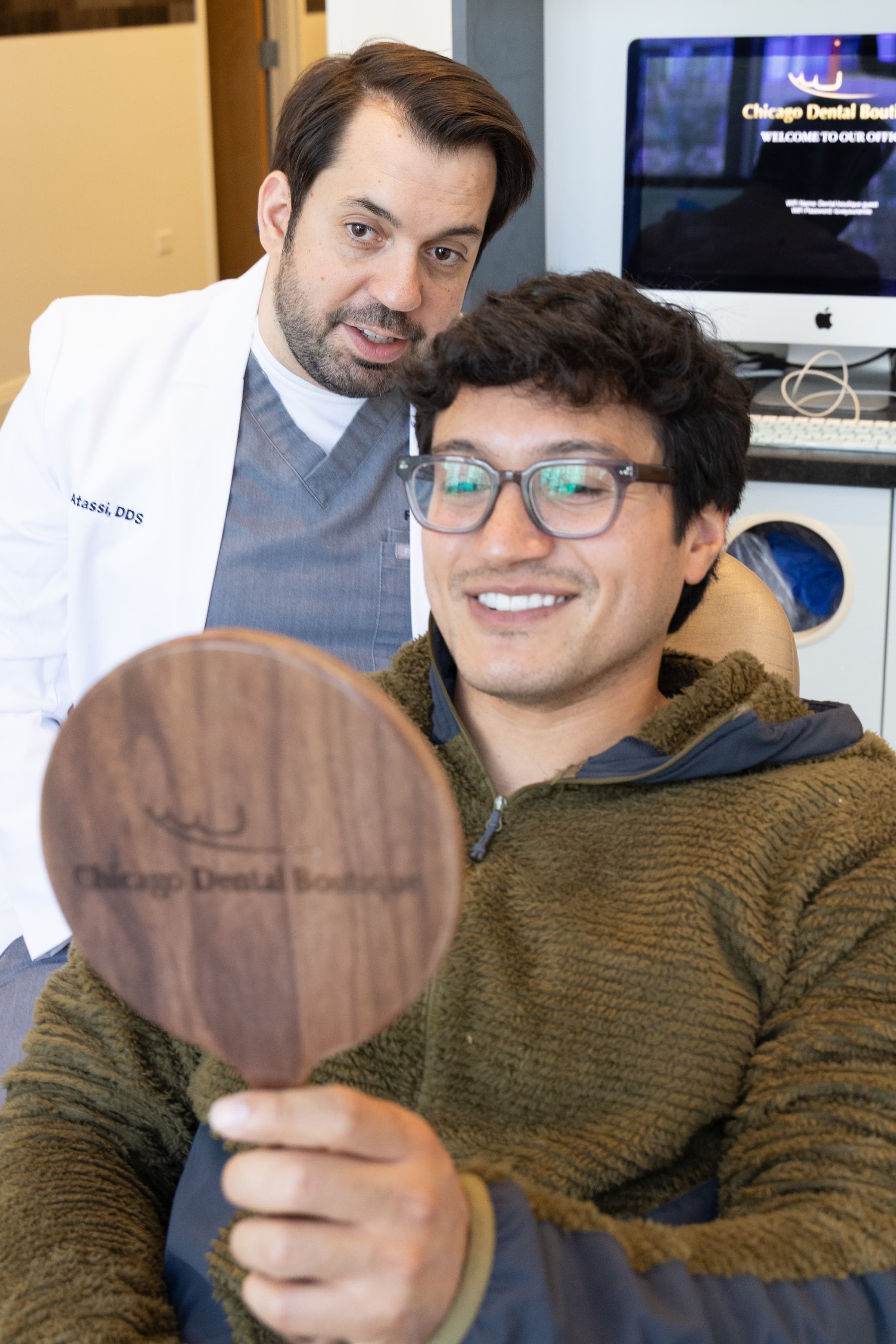 A patient smiling into a hand mirror beside Dr. Atassi
