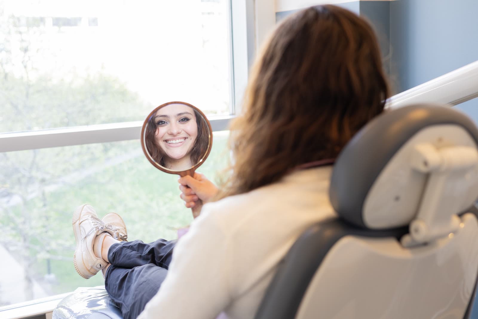 A patient looking at her smile in a hand mirror by the window